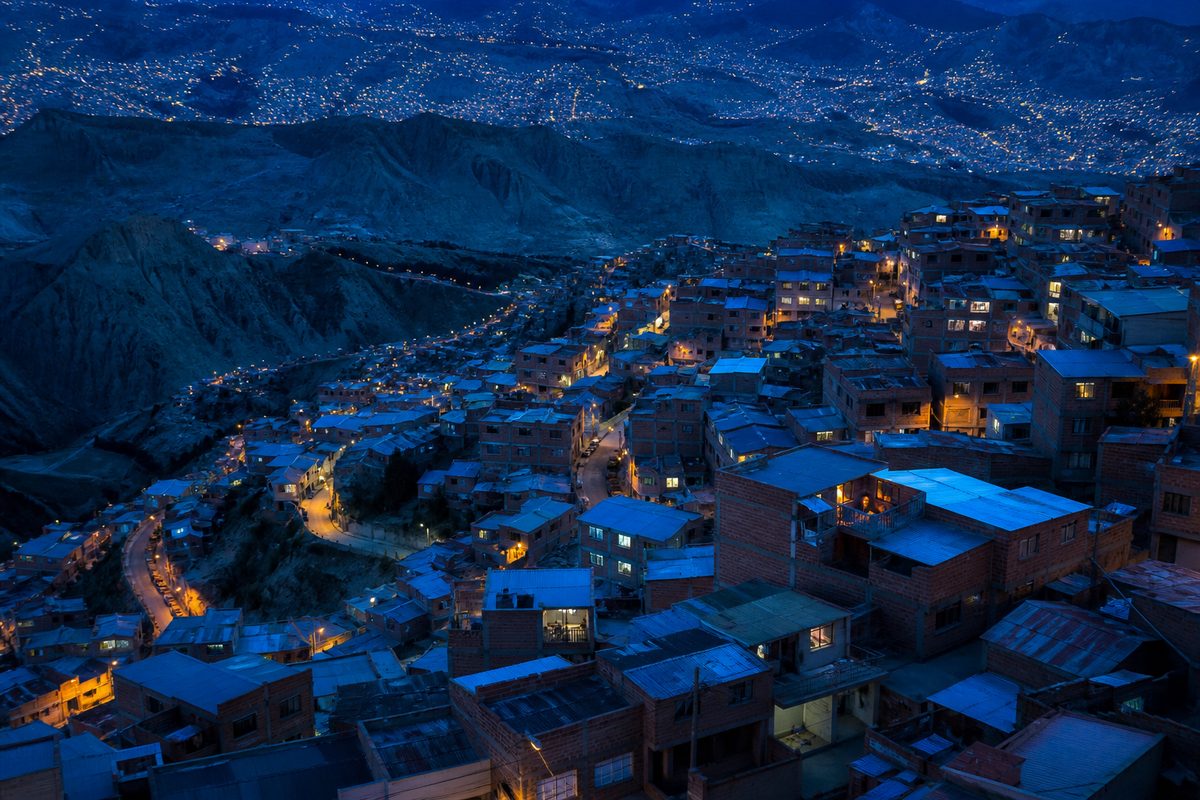 Vista aérea al anochecer de un barrio en ladera con casas apiladas y ventanas iluminadas